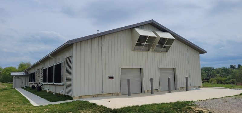 Long beige building with air handlers under blue sky green trees in the background.