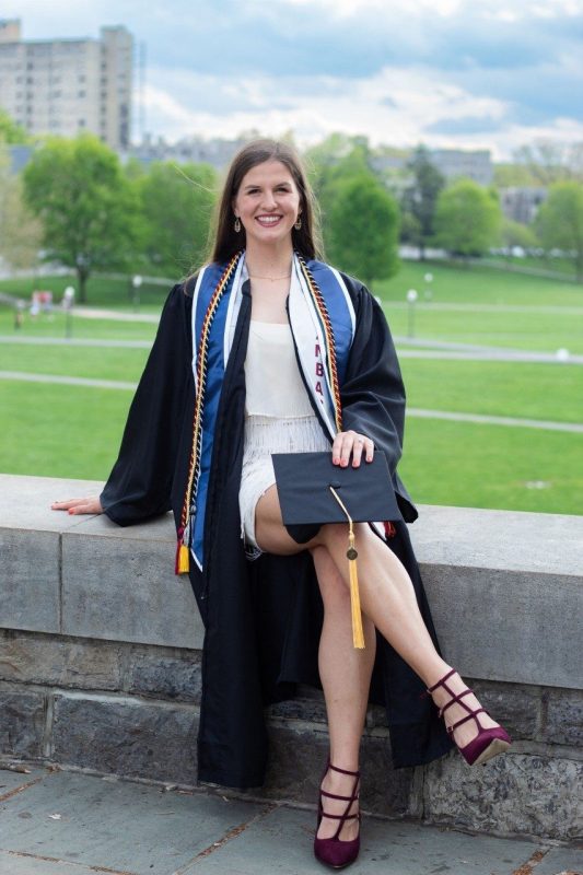 Outstanding senior for APSC, Katie Kirkpatrick in her graduation gown, cords, and stole, holding her cap, while seated with views of campus in the background.