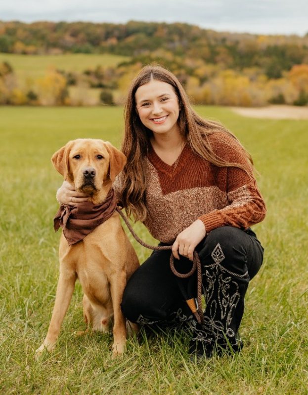White woman with long brown hair posing with her yellow lab dog in a green meadow. Dog is wearing a brown bandana, coordinating with the woman's sweater. 