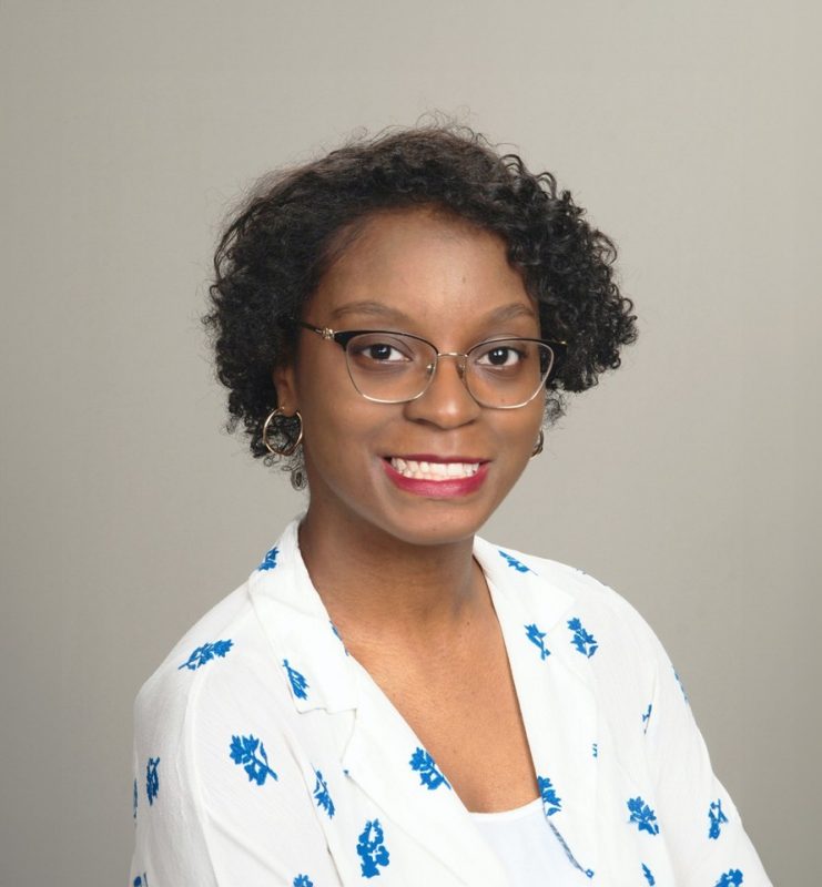Professional photo of a black woman with glasses wearing a with  jacket with blue pattern smiling into the camera.