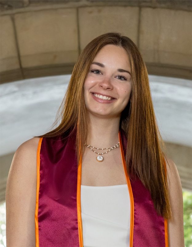 Graduate student, Camille Mitchell, smiling, wearing graduation sash in front of gothic arch on Virginia Tech campus.