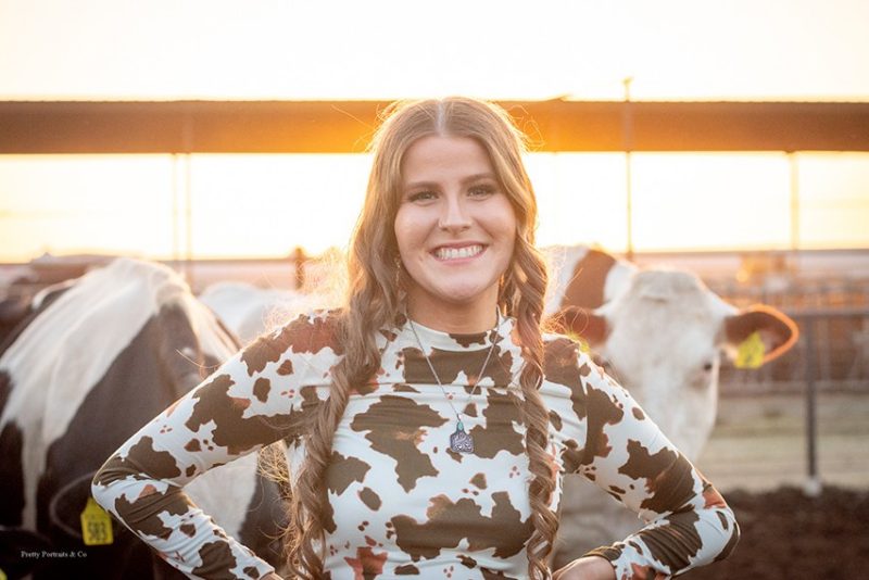 Graduate student, Holly Parks, wearing a cow print shirt with cattle in the background and sun on the horizon.
