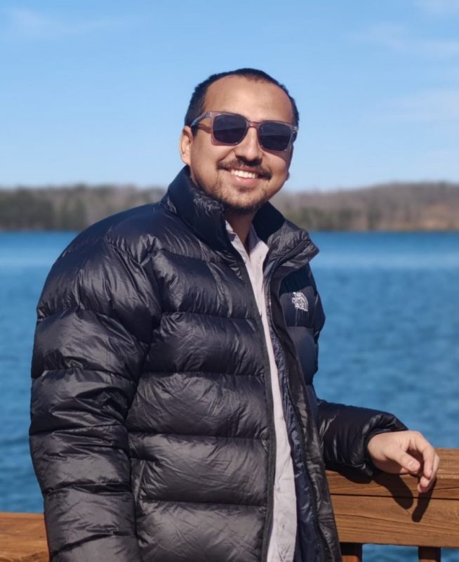 Photo of Binod Pokhrel in sunglasses and a parka, leaning against a wooden rail with a lake in the background and blue skies.