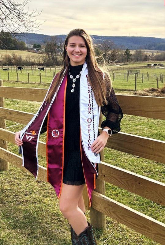 White woman with long brunette hair wearing a short black dress and black cowboy boots along with her Virginia Tech graduation sashes. Standing in front of a wood plank fence at the Virginia Tech farm with beef cattle in the pasture behind her and a view of the ridgetop in the background.