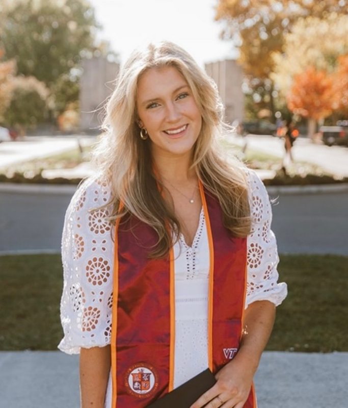 White woman with long blond hair smiling into the camera wearing a white dress with her Virginia Tech graduation sash.