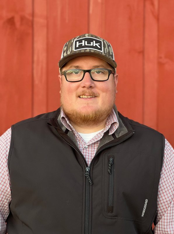 Bearded man with glasses, wearing a ball cap and black vest over a plaid button-down shirt in front of a red barn.