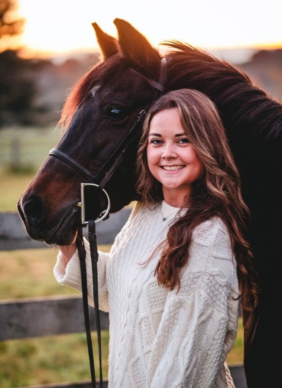 Young woman wih long hair with her horse at sunset. Wooden fence and pasture in the background.