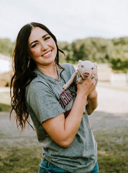 Smiling young woman with long dark hair wearing a gray Virginia Tech T-shirt holding a pink piglet.